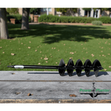Black 5" auger on weathered wooden board, with a blurred green lawn and trees in the background. 